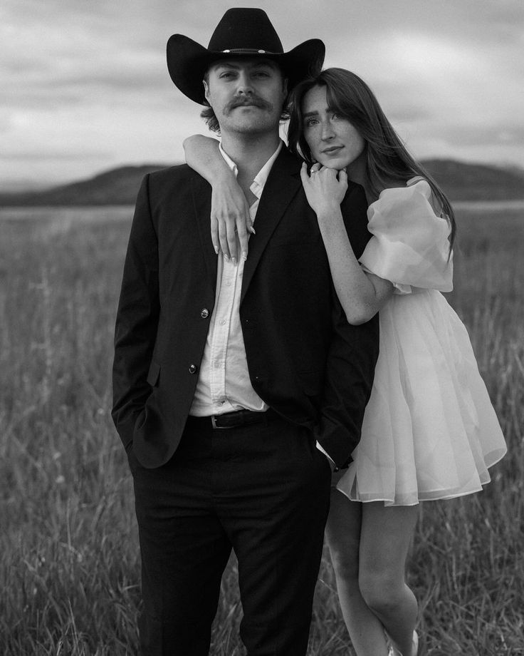 a man and woman are standing in the middle of a field with tall grass, one is wearing a cowboy hat