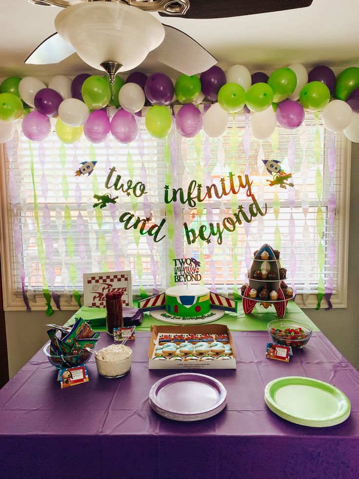 a purple table topped with lots of cake next to a window covered in green and white balloons