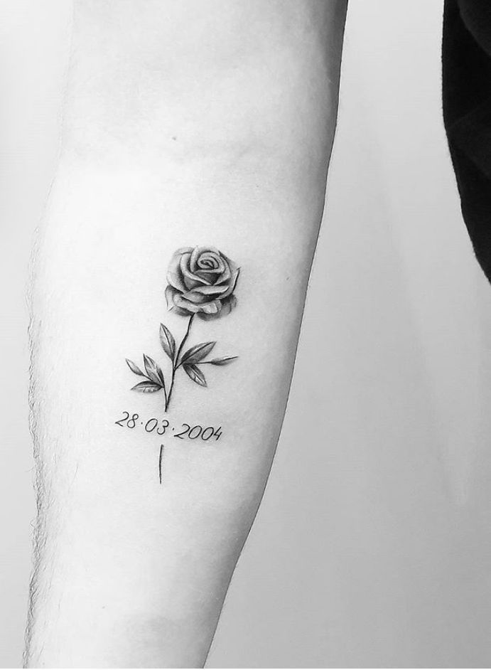 a black and white photo of a rose tattoo on the left arm, with date written in cursive writing