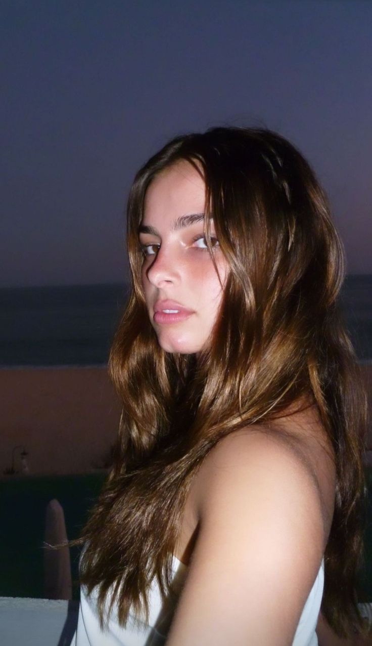 a woman with long brown hair standing next to the ocean at night looking off into the distance