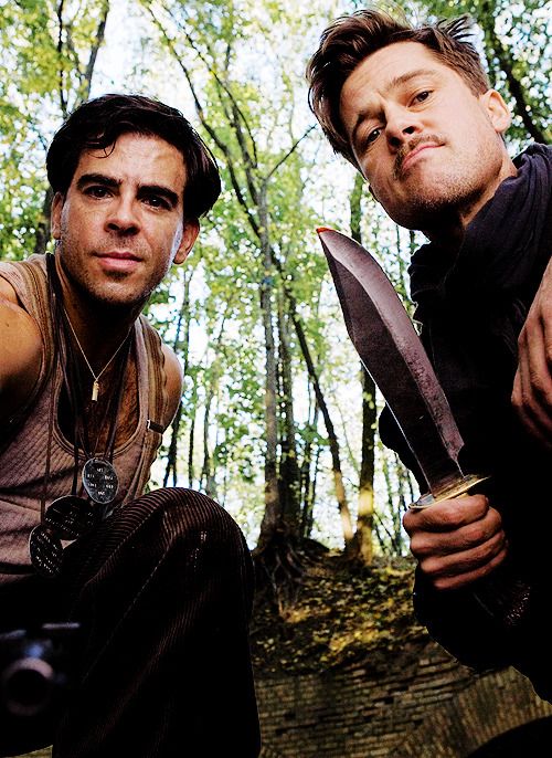 two men holding knives in the woods with one looking at the camera while the other looks up