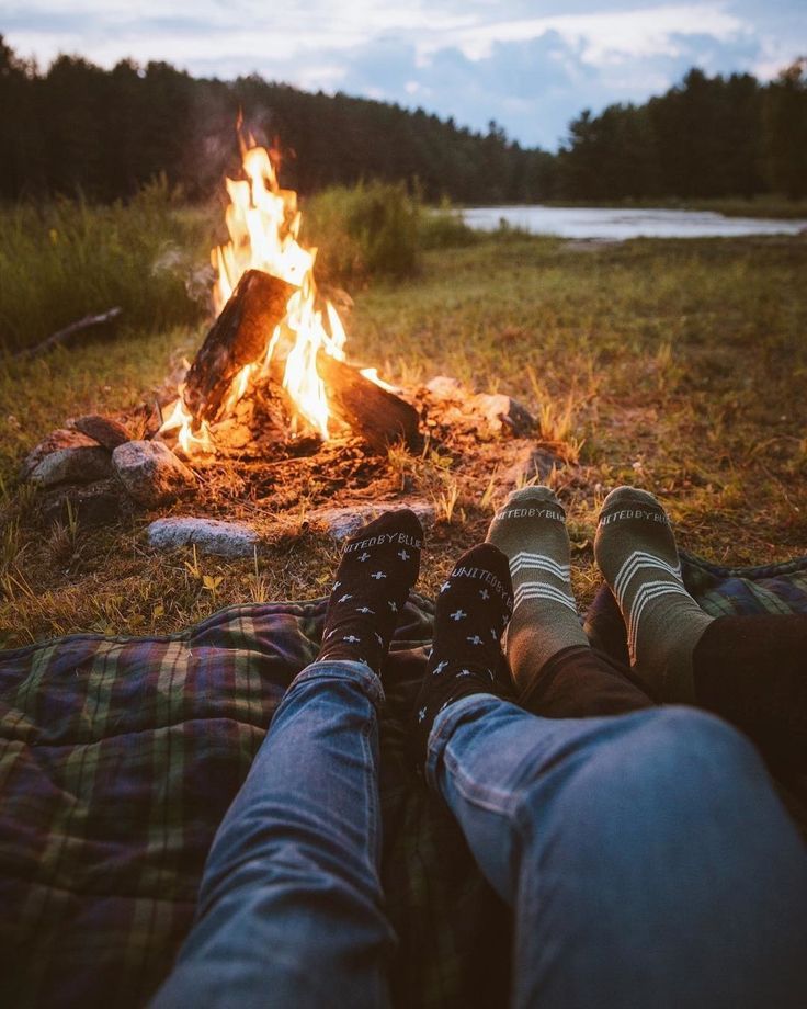 two people sitting in front of a campfire with their feet up on the ground