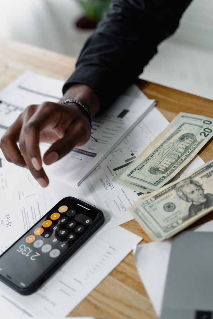 a person pointing at money on top of a desk with a calculator and pen