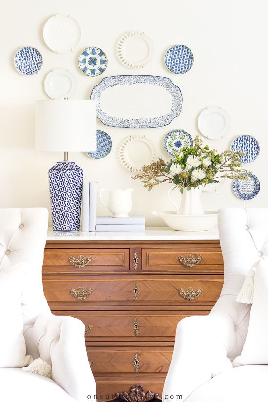 two white chairs sitting in front of a dresser with plates on the wall behind them