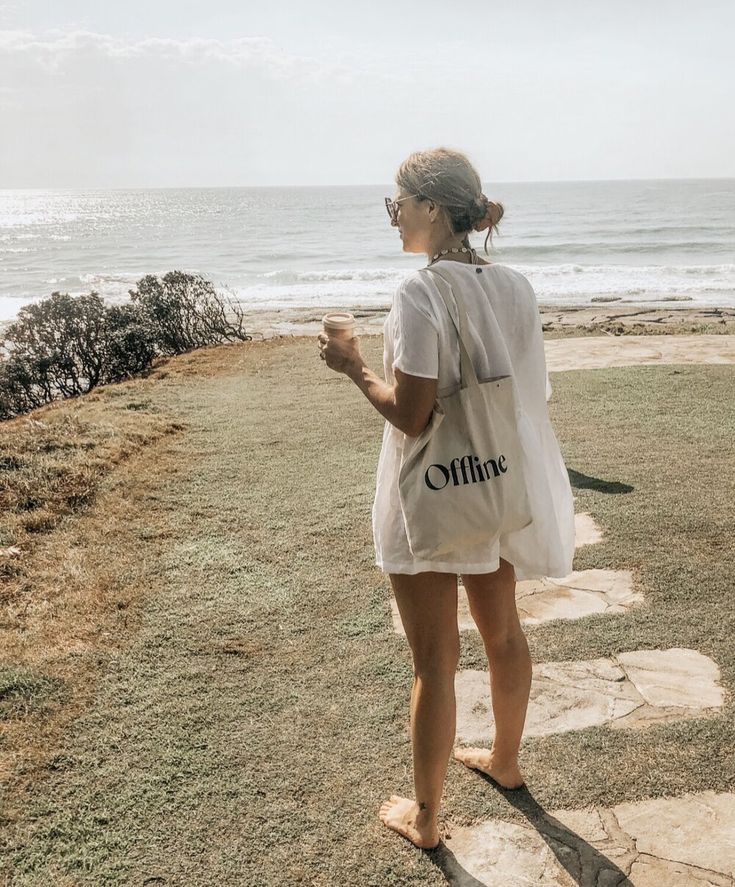 a woman standing on top of a grass covered field next to the ocean holding a coffee cup