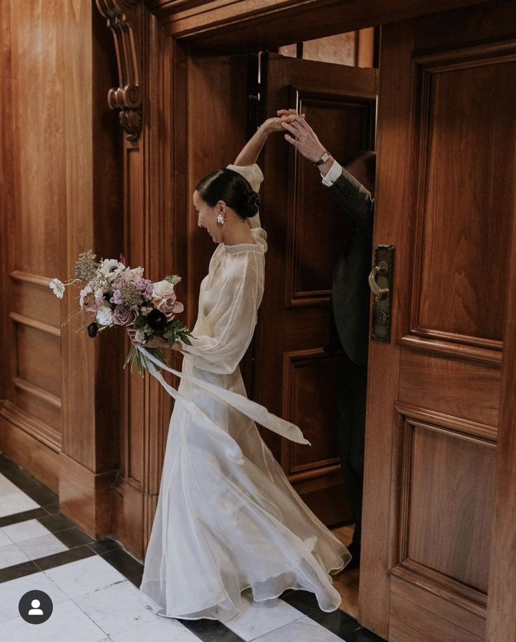 a woman in a white dress is standing near a door and holding a flower bouquet