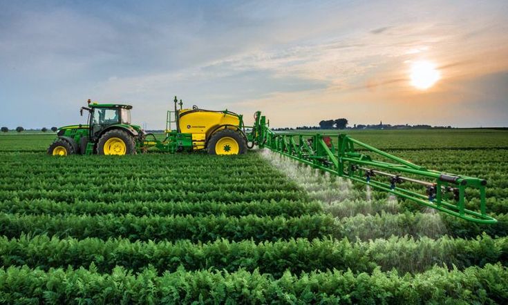 a tractor sprays pesticide on a green field at sunset with the sun in the background