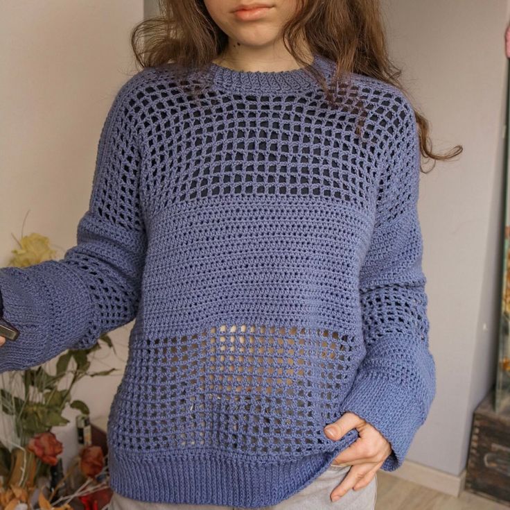 a woman wearing a blue sweater with holes in the front and back, standing next to a plant