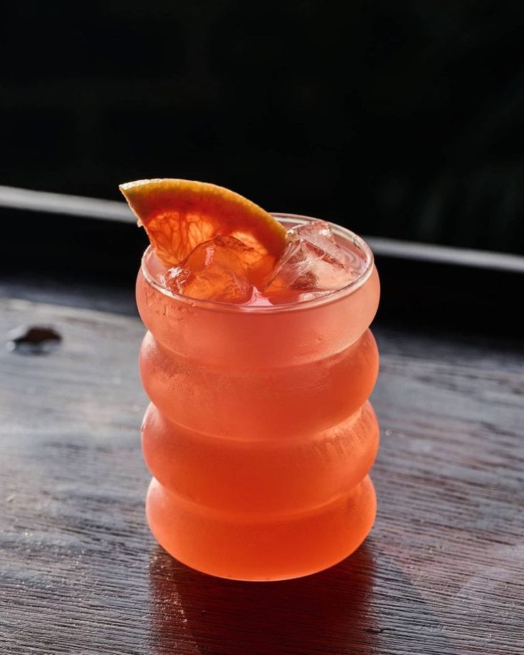 an orange drink in a tall glass on a wooden table with a slice of fruit