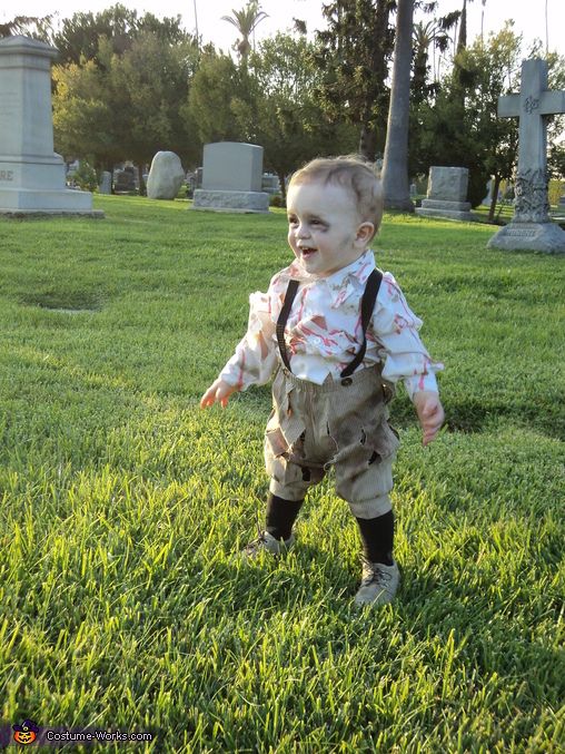 a toddler dressed in zombie clothes walking through the grass at a cemetery with his mouth open