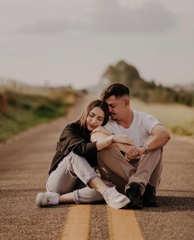 a man and woman sitting on the side of an empty road with their arms around each other