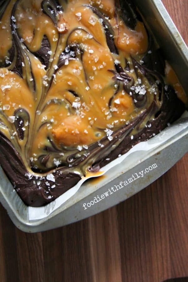 a cake in a pan with chocolate and caramel toppings on the top, sitting on a wooden table