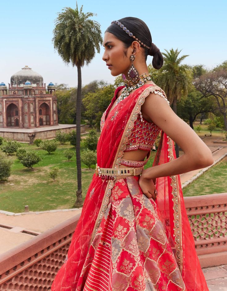 This exquisite banarasi lehenga features a peek-through panel that adds a contemporary touch to the ogee pattern, adorned with delicate floral motifs for a stunning overall design. Paired with an embroidered V-neck blouse, it is complemented by a light organza side-draped dupatta, enhancing the elegance of the ensemble. The look is beautifully finished with our signature AG belt, adding a refined touch to this captivating piece. Fitted Pink Lehenga With Bandhani Print, Pink Bandhani Print Semi-stitched Lehenga, Pink Floral Print Lehenga With Traditional Drape, Festive Pink Lehenga With Cutdana Detail, Unstitched Pink Lehenga With Cutdana Details, Ogee Pattern, Silk Lehenga, Organza Dupatta, V Neck Blouse