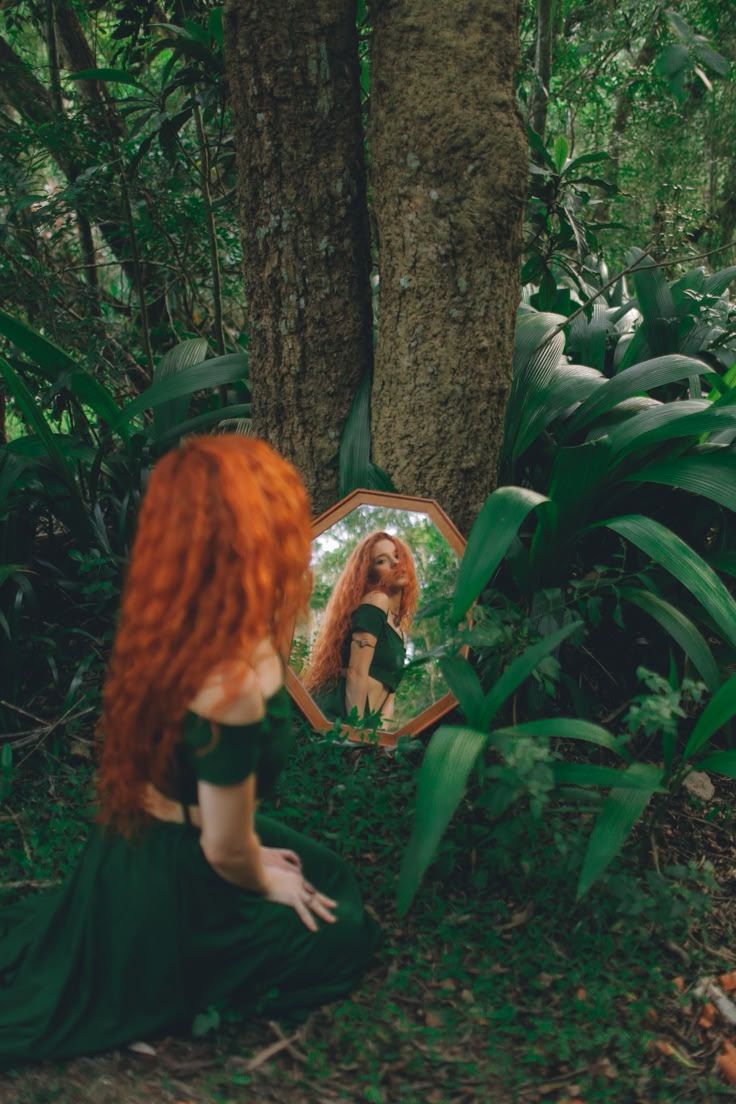 a woman with red hair sitting in the woods looking at her reflection in a mirror