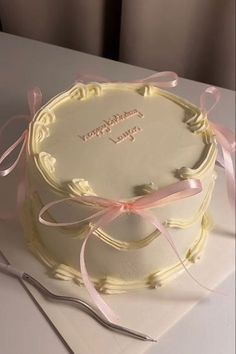 a white cake with pink ribbon on top sitting on a table next to a fork and knife