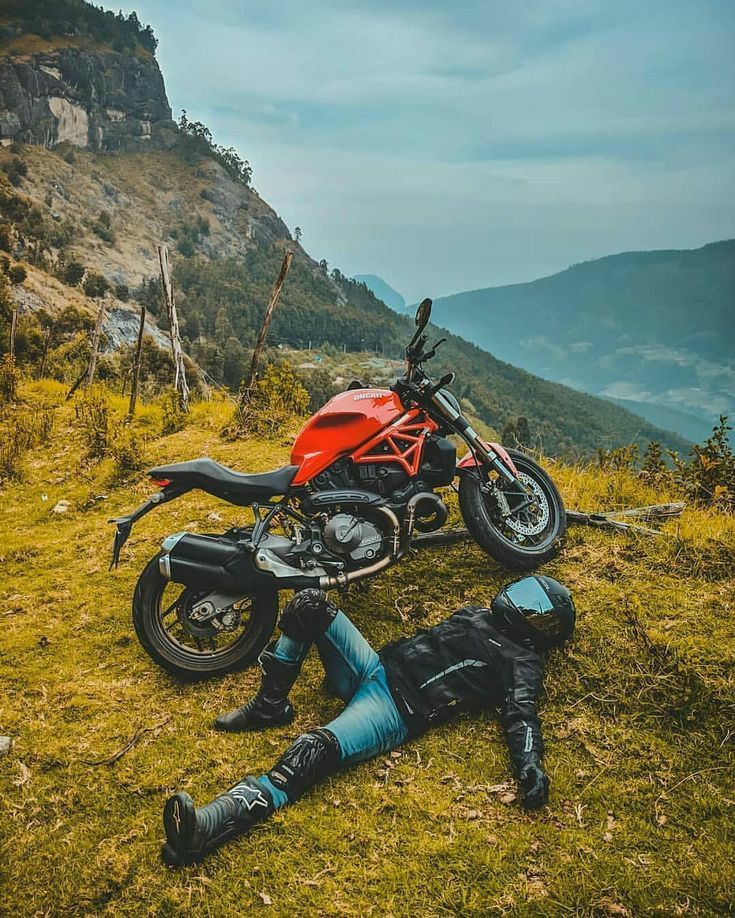 a red motorcycle parked on top of a lush green hillside next to a person laying on the ground