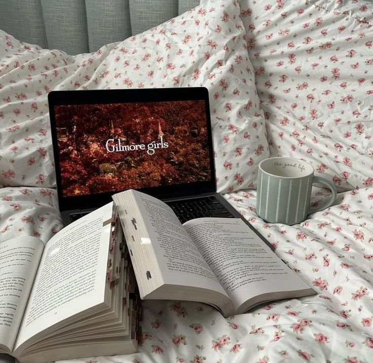 an open laptop computer sitting on top of a bed next to two books and a cup