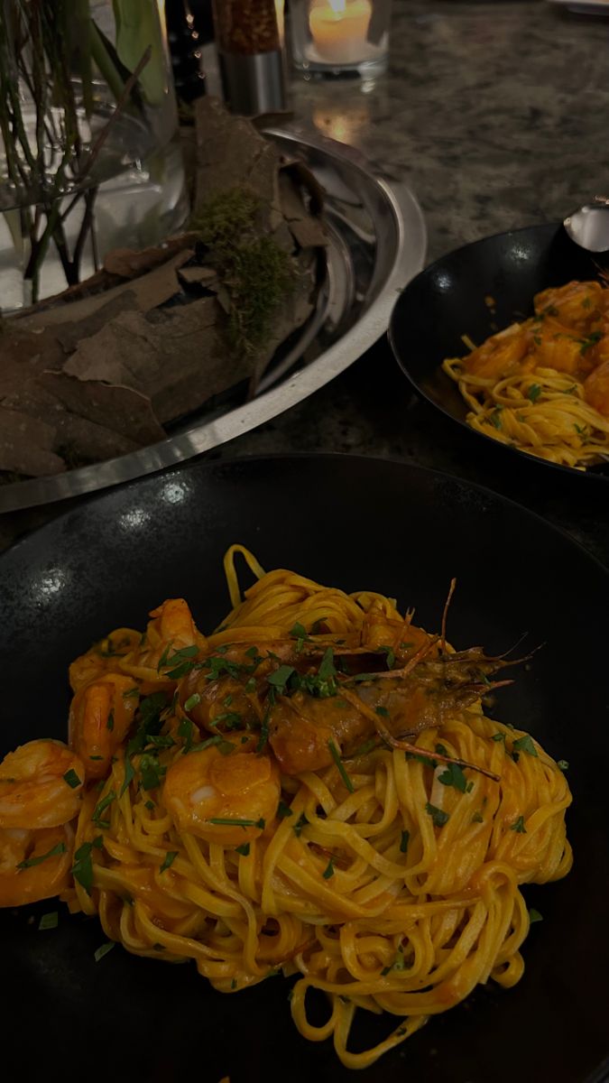 two black plates filled with pasta and shrimp on top of a table next to candles