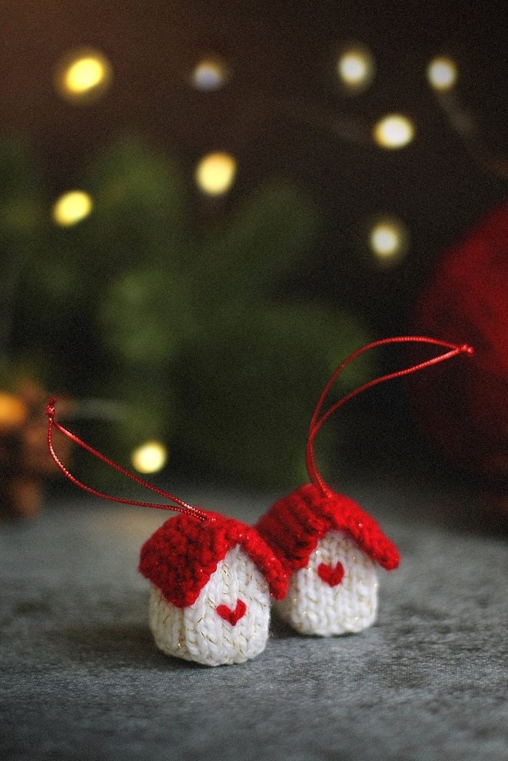 two small crocheted houses hanging from strings on a table with christmas lights in the background