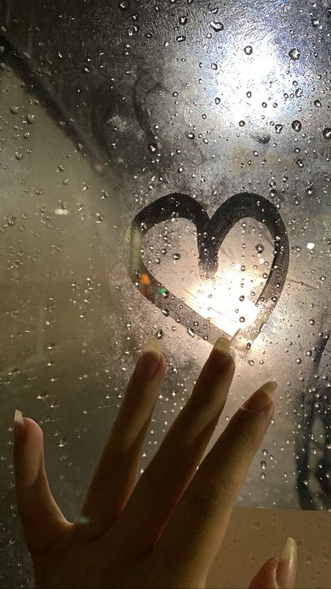 a person's hand holding up a heart shaped object through a window with rain drops on it