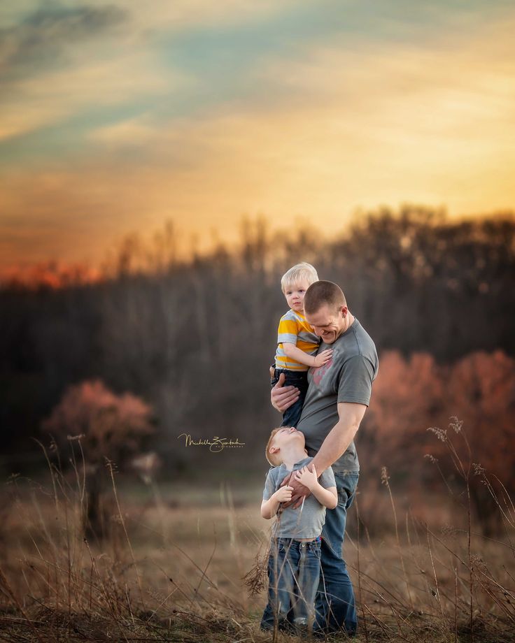 a man holding a baby in his arms while standing in a field with trees behind him