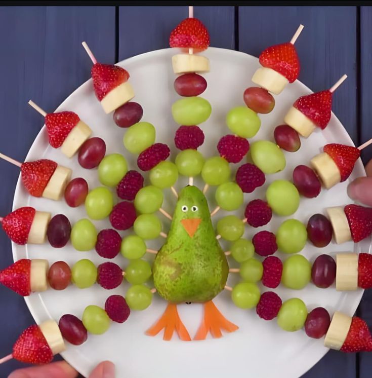 a white plate topped with fruit and veggies next to a hand holding a green bird