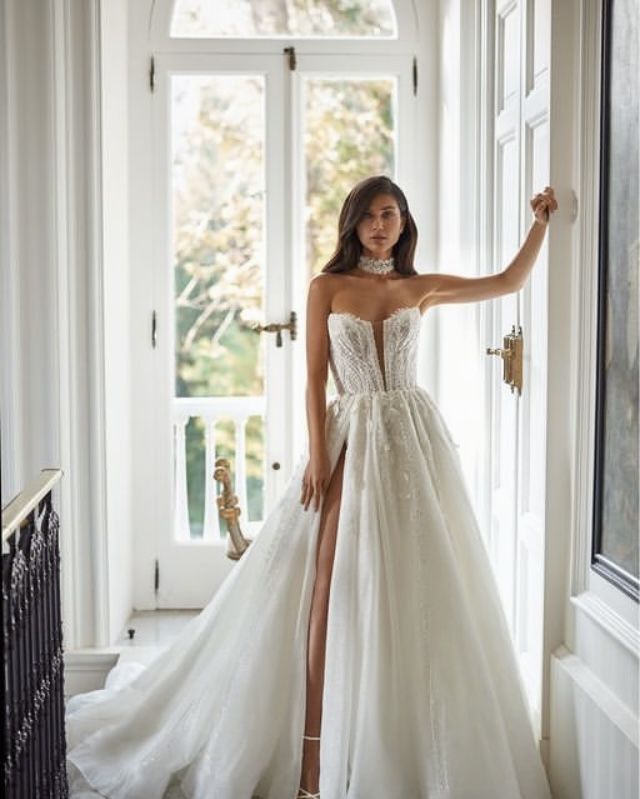 a woman standing in front of a window wearing a wedding dress with high slits