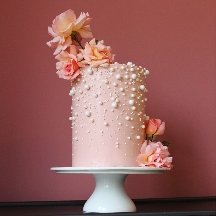 a pink cake with pearls and flowers on top is sitting on a white pedestal in front of a pink wall