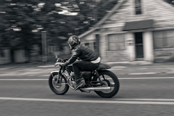 black and white photograph of a person riding a motorcycle down the street with houses in the background