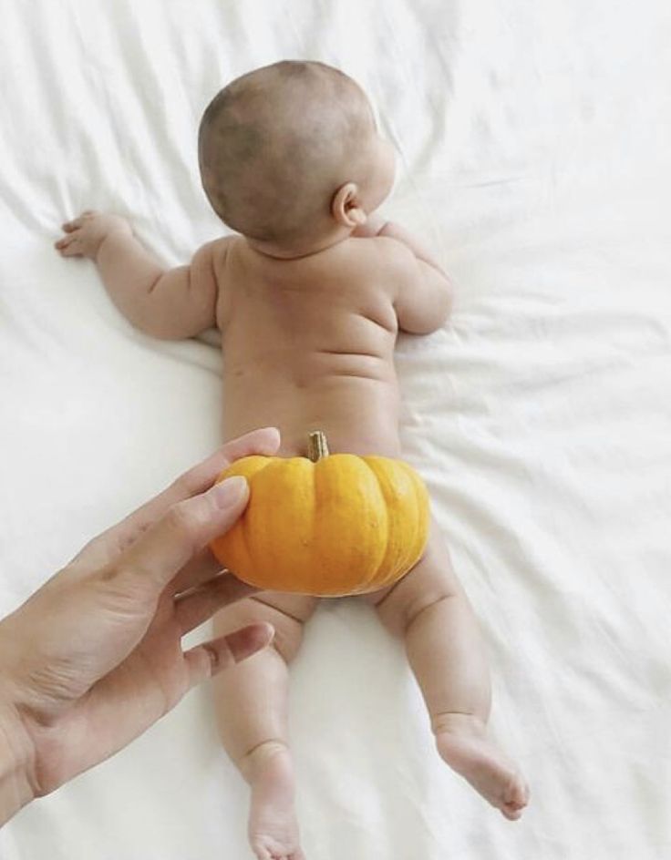 a baby laying on top of a bed next to a person holding a small pumpkin