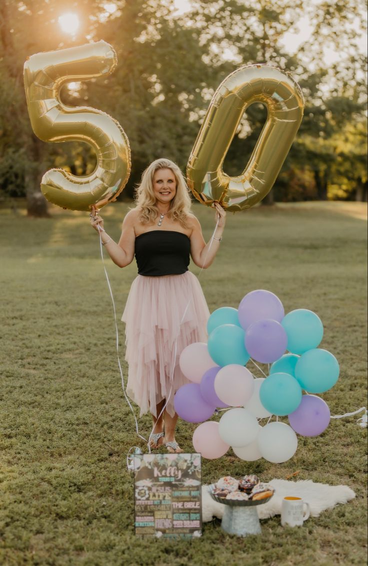 a woman in a dress holding balloons and posing for the camera with her 50th birthday cake