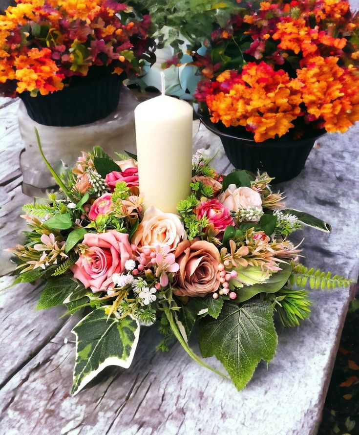 an arrangement of flowers on a table with a candle in the center and potted plants behind it