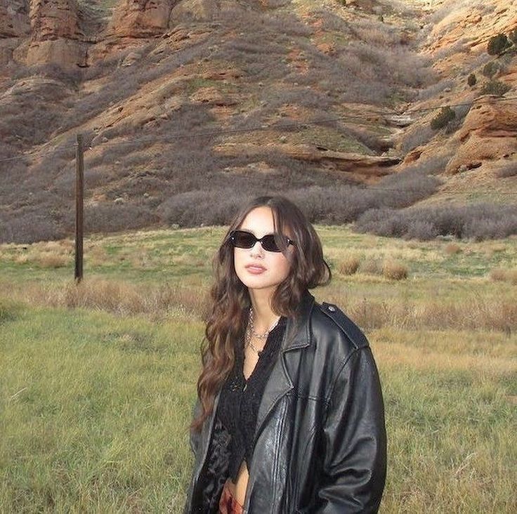 a woman wearing sunglasses standing in front of a mountain with grass and rocks behind her