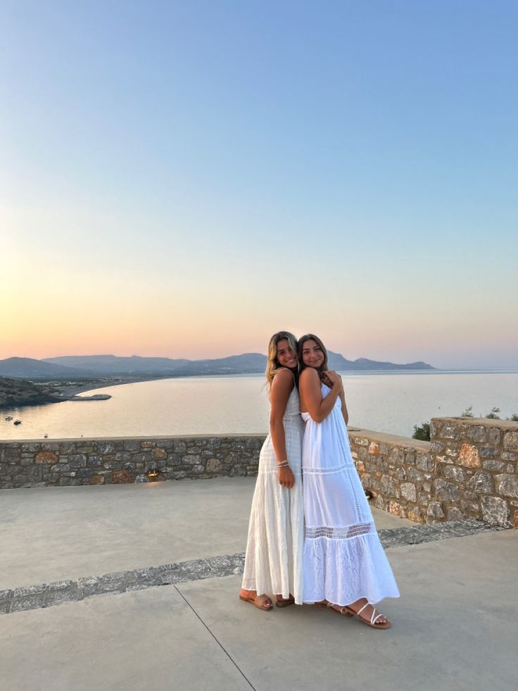 two women standing next to each other in front of a body of water at sunset