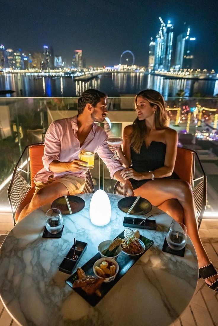 two people sitting at a table with food and drinks in front of the city skyline