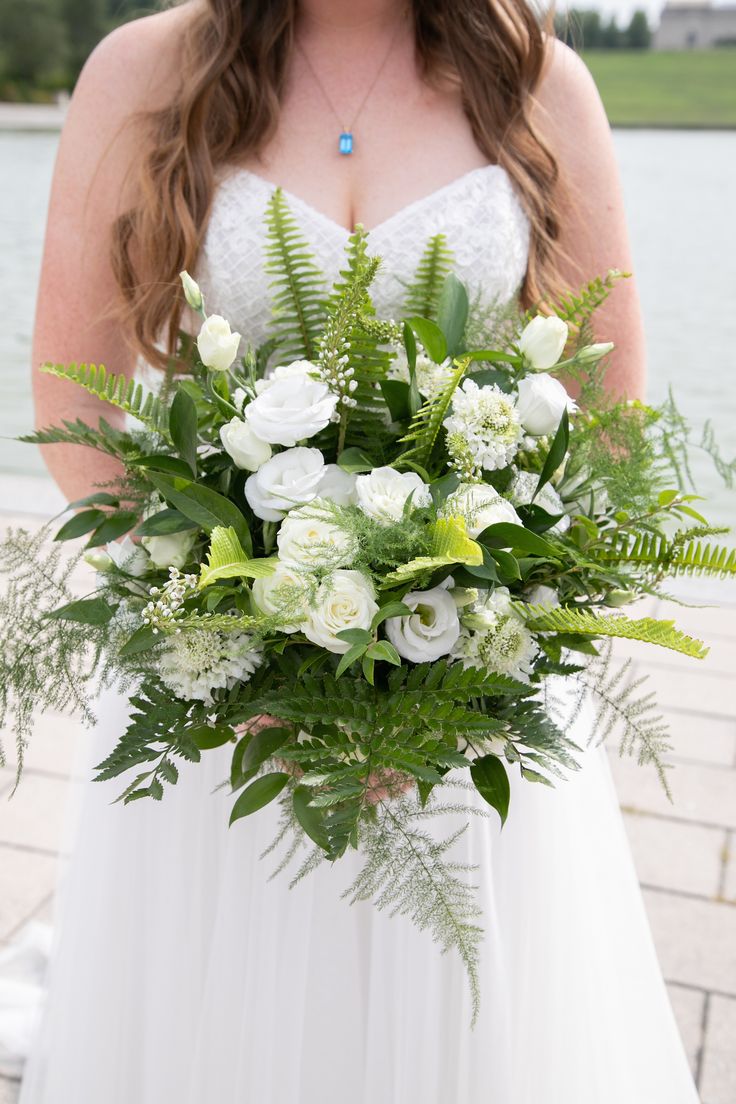 a bride holding a bouquet of white flowers and greenery in front of a lake