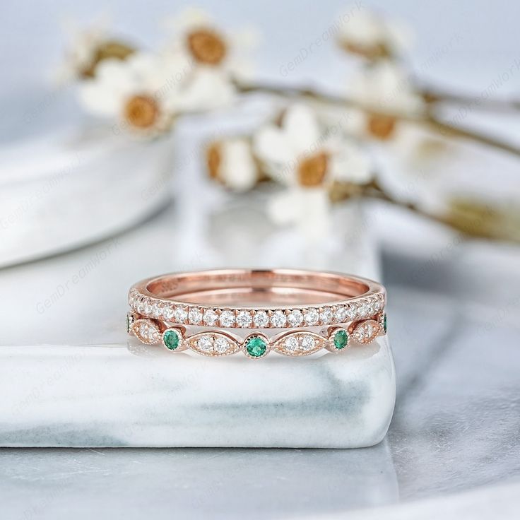 two wedding bands with emeralds and diamonds on top of a white marble table next to flowers