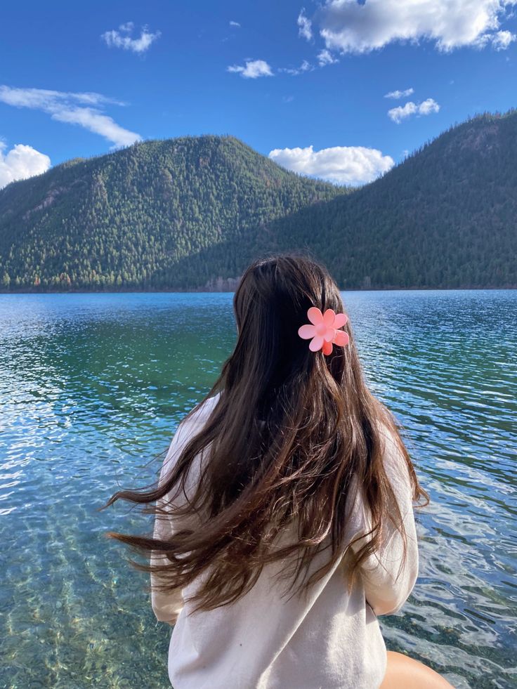 a woman sitting on the edge of a body of water with a pink flower in her hair