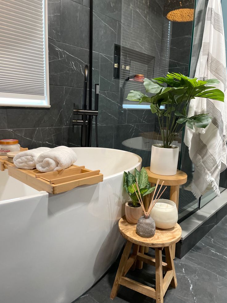 a white bath tub sitting inside of a bathroom next to a wooden stool and potted plant