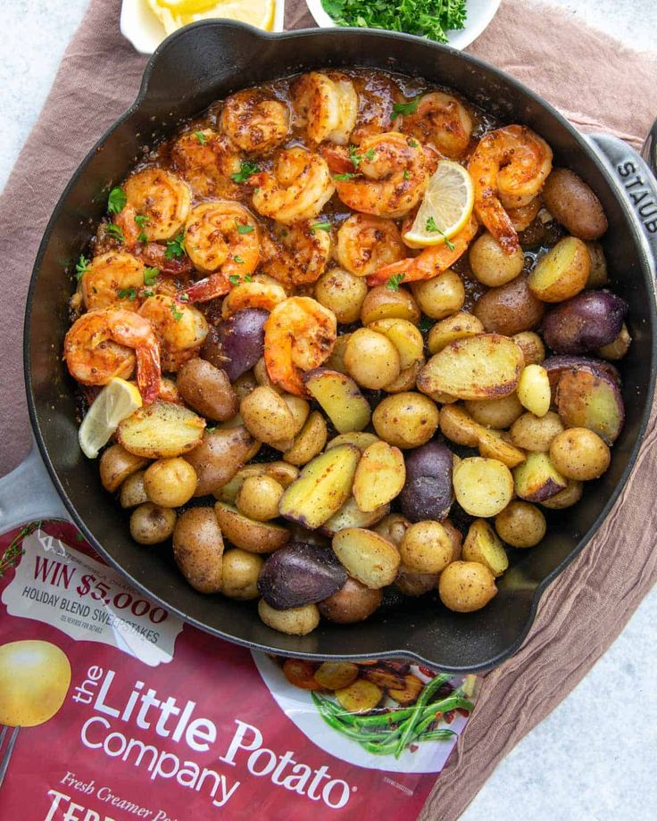 a pan filled with potatoes and shrimp on top of a table next to lemon wedges