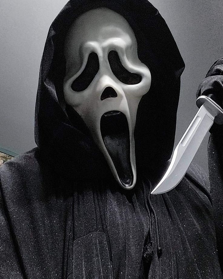 a person in a black hooded jacket with a white mask on holding a knife up to their face