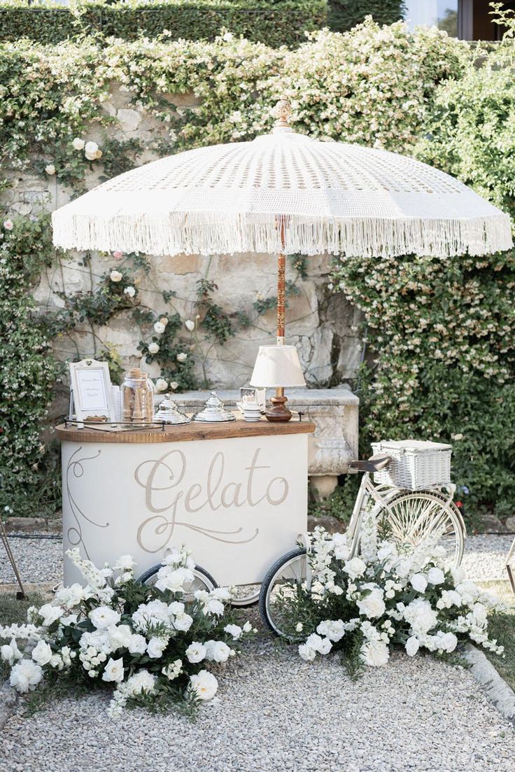 an ice cream stand with white flowers and umbrella in front of a stone wall that says gelato