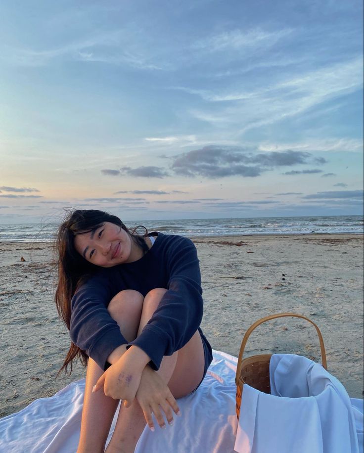 a woman sitting on top of a white blanket next to the ocean with her arms crossed