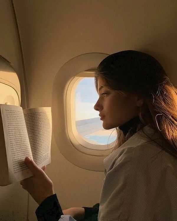 a woman reading a book while sitting on an airplane with the window looking out at the ocean