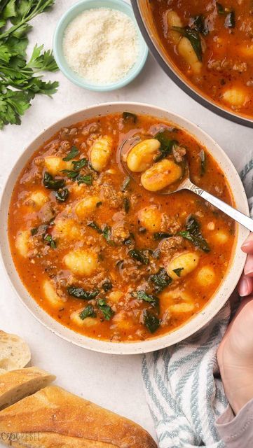 a person holding a spoon in a bowl of stew with bread and parsley on the side