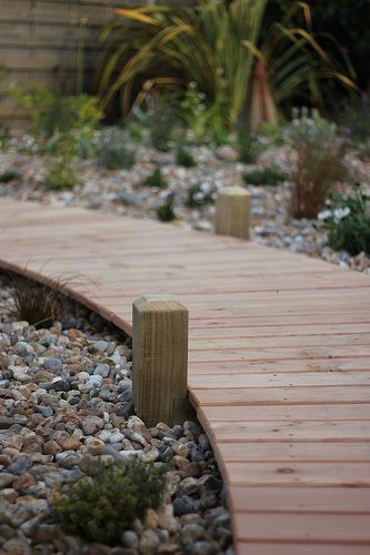 a wooden walkway surrounded by gravel and rocks in a garden area with plants on either side