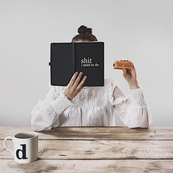 a woman sitting at a table reading a book and holding a slice of pizza in front of her face