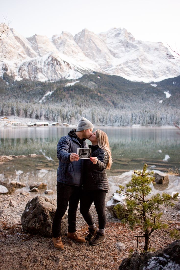 a man and woman standing next to each other in front of a lake with mountains