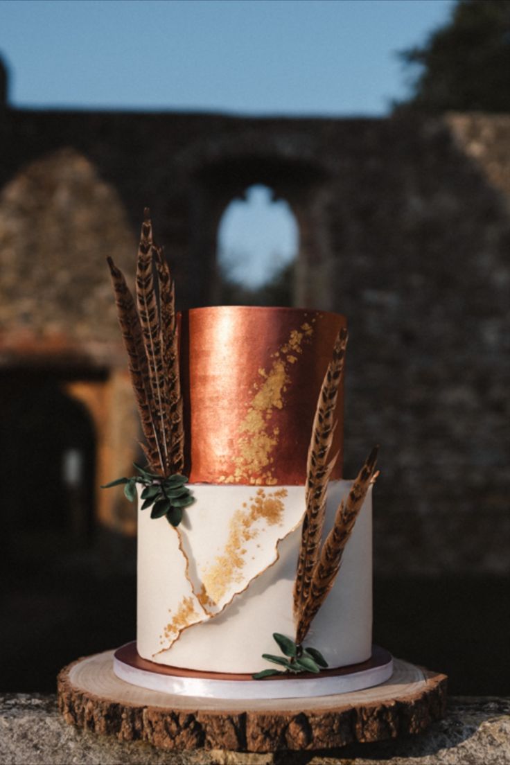 a white and gold wedding cake with feathers on the top is sitting on a tree stump