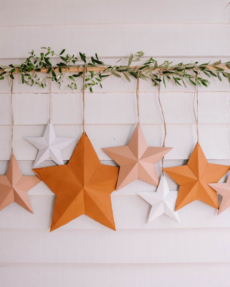 five paper stars hanging on a wall with greenery in the corner and one is orange